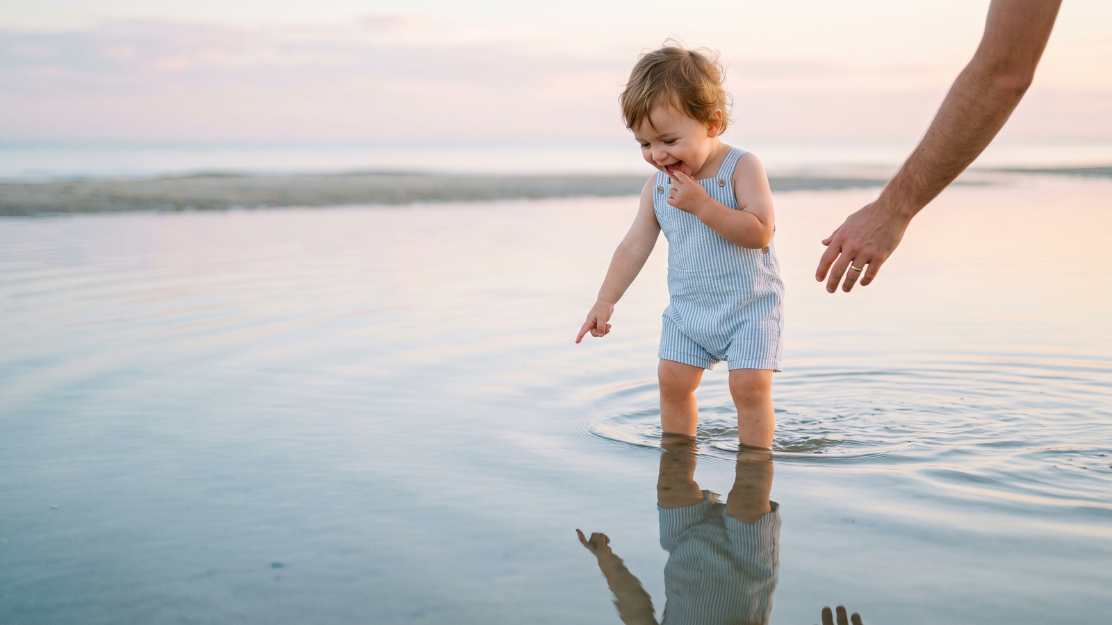 Photo of a toddler ankle deep in glassy calm lagoon water reaching toward their reflection with a parent's hand near